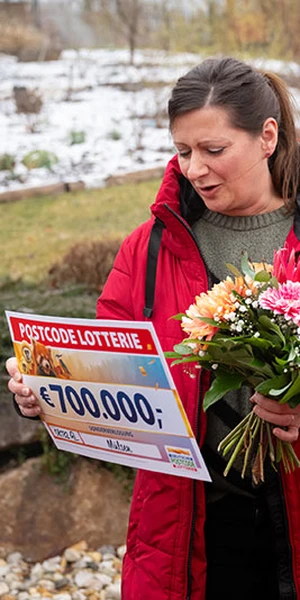 Eine Frau in rotem Mantel hält einen Blumenstrauß und einen großen Scheck über 700.000 € von der Postcode Lotterie. Sie steht im Freien vor einer verschneiten Kulisse.