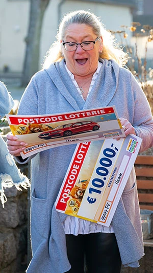 Eine überraschte Frau hält einen großen Postcode-Lotterie-Scheck über 10.000 € in den Händen. Sie steht im Freien in einem ungezwungenen Umfeld.