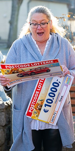 Eine überraschte Frau hält einen großen Postcode-Lotterie-Scheck über 10.000 € in den Händen. Sie steht im Freien in einem ungezwungenen Umfeld.