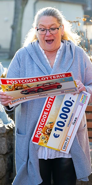 Eine überraschte Frau hält einen großen Postcode-Lotterie-Scheck über 10.000 € in den Händen. Sie steht im Freien in einem ungezwungenen Umfeld.
