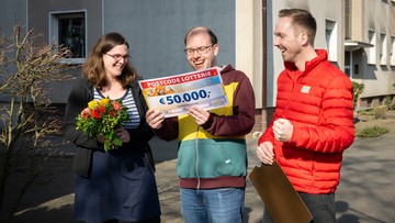 Drei Personen feiern im Freien. Eine von ihnen hält einen großen Scheck über 50.000 € von der Postcode Lotterie, eine andere trägt einen Blumenstrauß.