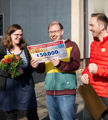 Drei Personen feiern im Freien. Eine von ihnen hält einen großen Scheck über 50.000 € von der Postcode Lotterie, eine andere trägt einen Blumenstrauß.