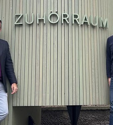 Zwei Männer stehen vor einem Gebäude mit einem Schild, auf dem „Zuhörraum“ steht. Einer trägt ein Sakko, der andere einen Pullover. In der Nähe befindet sich eine bunte Bank.