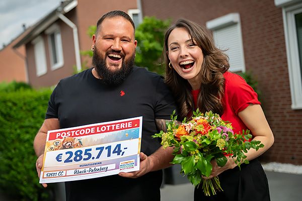 Ein Mann hält einen Scheck der Deutschen Postcode Lotterie in den Händen. Eine Frau hält einen Blumenstrauß. Beide strahlen über beide Ohren.