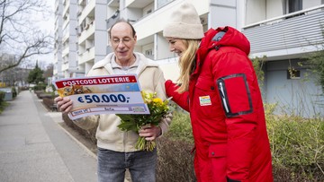 Ein Mann hält einen Lotterie-Scheck über 50.000 € und einen Blumenstrauß in den Händen und lächelt. Neben ihm steht eine Frau in rotem Mantel und Mütze. Sie befinden sich vor einem Mehrfamilienhaus.