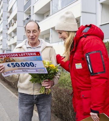 Ein Mann hält einen Lotterie-Scheck über 50.000 € und einen Blumenstrauß in den Händen und lächelt. Neben ihm steht eine Frau in rotem Mantel und Mütze. Sie befinden sich vor einem Mehrfamilienhaus.