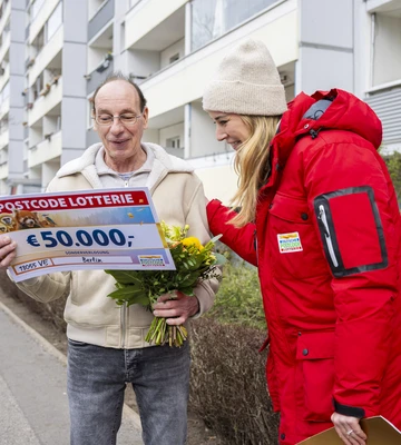 Ein Mann hält einen Lotterie-Scheck über 50.000 € und einen Blumenstrauß in den Händen und lächelt. Neben ihm steht eine Frau in rotem Mantel und Mütze. Sie befinden sich vor einem Mehrfamilienhaus.