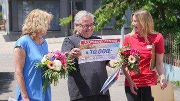 Three people smiling outdoors, holding large checks and flower bouquets. The checks display a €10,000 prize from a lottery.