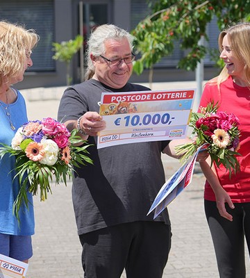 Three people smiling outdoors, holding large checks and flower bouquets. The checks display a €10,000 prize from a lottery.