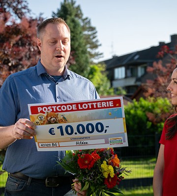 Man holding a large check for €10,000 from Postcode Lottery, standing next to a woman with flowers, in a garden setting.
