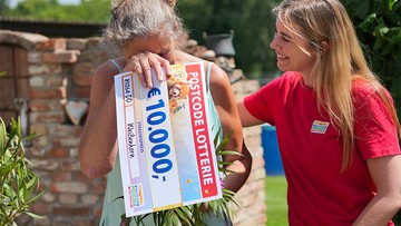 A woman joyfully hides her face behind a large 10,000 Euro lottery check, while another woman in a red shirt smiles beside her outdoors.