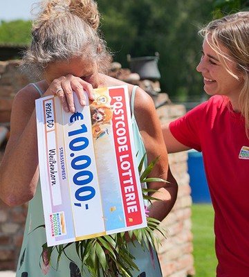 A woman joyfully hides her face behind a large 10,000 Euro lottery check, while another woman in a red shirt smiles beside her outdoors.