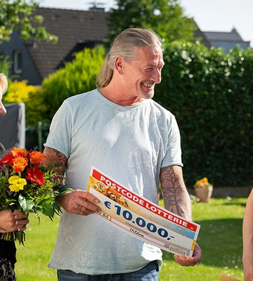 A smiling man holds a €10,000 lottery check, standing with two women; one holds a bouquet. They are outdoors in a garden setting.