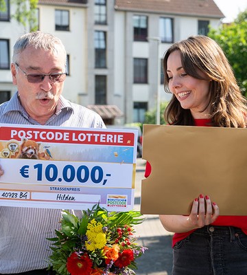 Two people smiling, holding a large lottery check for €10,000 and a bouquet of flowers, standing outside with houses in the background.