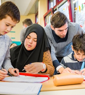 Eine Lehrkraft hilft drei jungen Schüler*innen bei ihren Schulaufgaben an einem Klassentisch, wobei der Schwerpunkt auf Schreibübungen liegt.