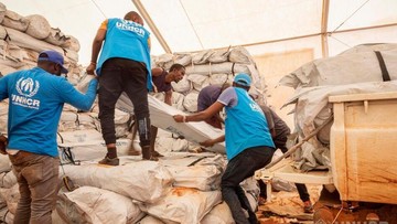 UNHCR-Mitarbeiter in blauen Westen entladen Hilfsgüter von einem Lastwagen und stapeln sie in einem Zelt.