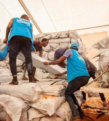 UNHCR-Mitarbeiter in blauen Westen entladen Hilfsgüter von einem Lastwagen und stapeln sie in einem Zelt.