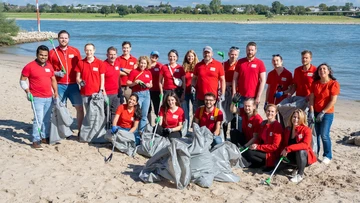 A group of people in red shirts pose with trash bags and pickers on a sandy beach by a river, participating in a cleanup activity.