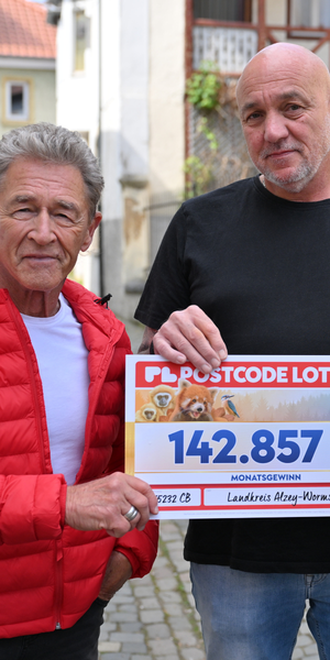 Zwei Männer stehen auf einer Kopfsteinpflasterstraße und halten einen Scheck der Postcode Lotterie über 142.857 Euro.