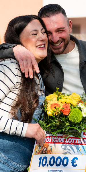 Fröhliches Paar umarmt sich draußen, hält einen Blumenstrauß und einen 10.000-Euro-Scheck der Postcode Lotterie und lächelt.