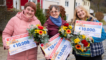 Drei lächelnde Frauen halten große 10.000-Euro-Schecks der Postcode Lotterie sowie bunte Blumensträuße.