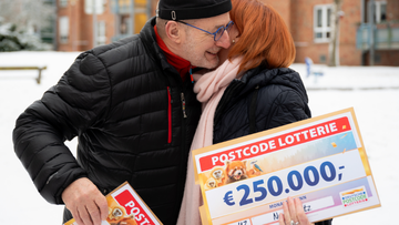 Zwei Personen umarmen sich freudig im Schnee und halten einen großen 250.000-Euro-Scheck der Postcode Lotterie.