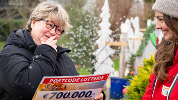 Frau im Mantel hält überrascht und glücklich einen 700.000-Euro-Lottoscheck, neben ihr steht eine weitere lächelnde Frau, draußen in einer verschneiten Umgebung.