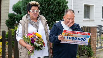 Überraschter Mann hält draußen einen 1.000.000-Euro-Lottoscheck, neben ihm steht eine Frau mit einem bunten Blumenstrauß.